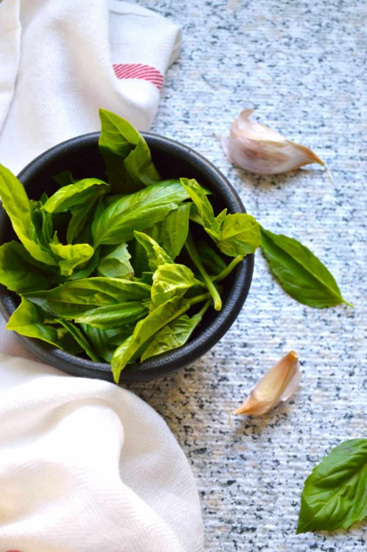 A black bowl of thai basil leaves with garlic cloves on a table.